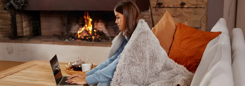 Γυναίκα εργάζεται στον υπολογιστή της, τυλιγμένη με κουβέρτα δίπλα σε αναμμένο τζάκι, απολαμβάνοντας οικονομική και άνετη θέρμανση στο σπίτι.